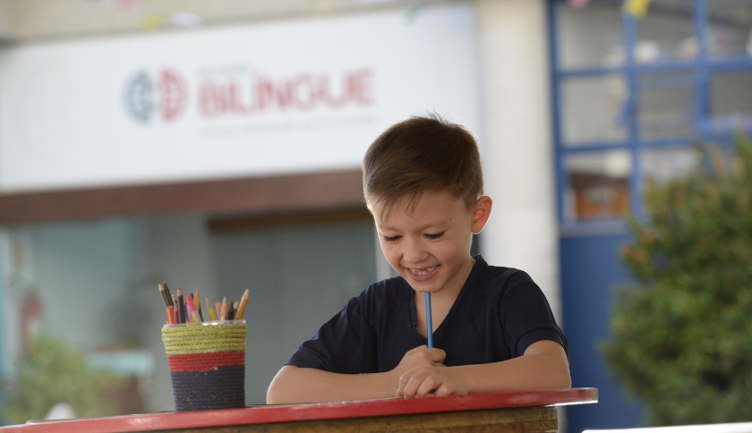 Criança desenhando e, ao fundo, a escola portal bilíngue